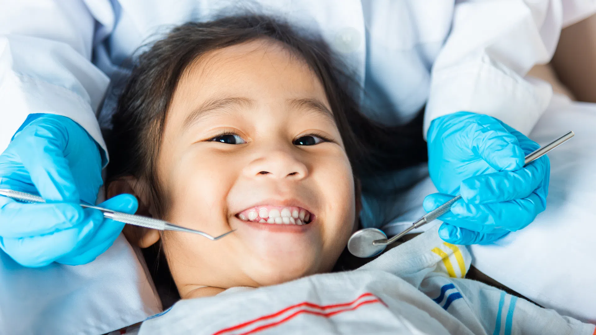 Smiling child receiving dental checkup with dentist wearing blue gloves and holding dental tools.