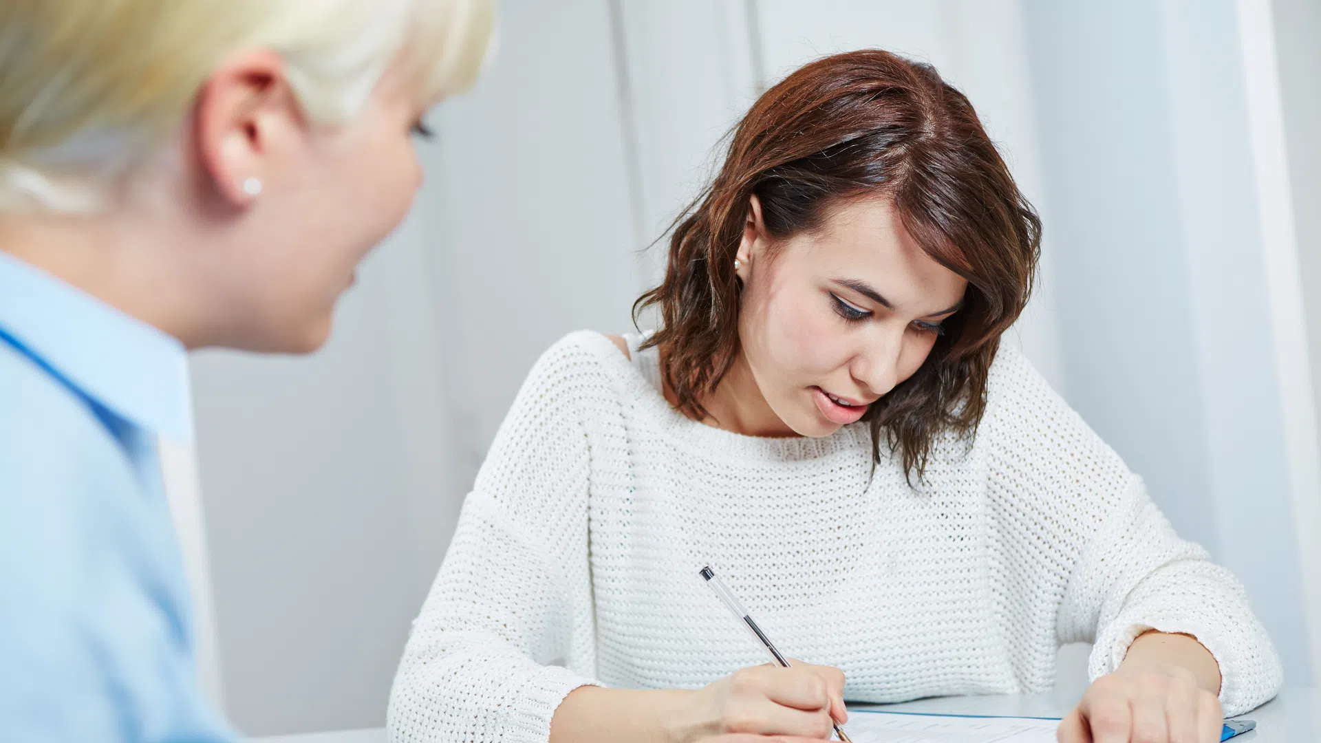 Young woman in white sweater signing documents while a person in blue shirt assists her at a table