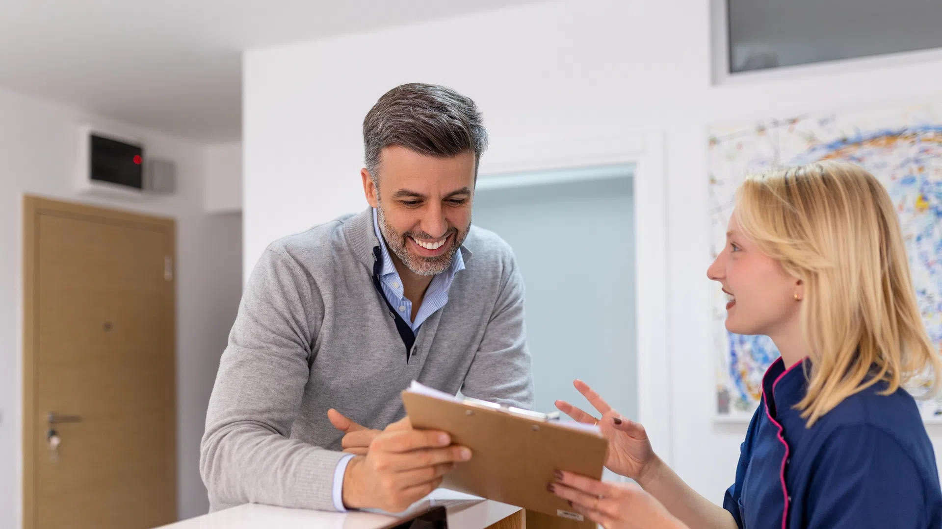 Smiling receptionist assisting male guest at hotel front desk with clipboard and registration form.