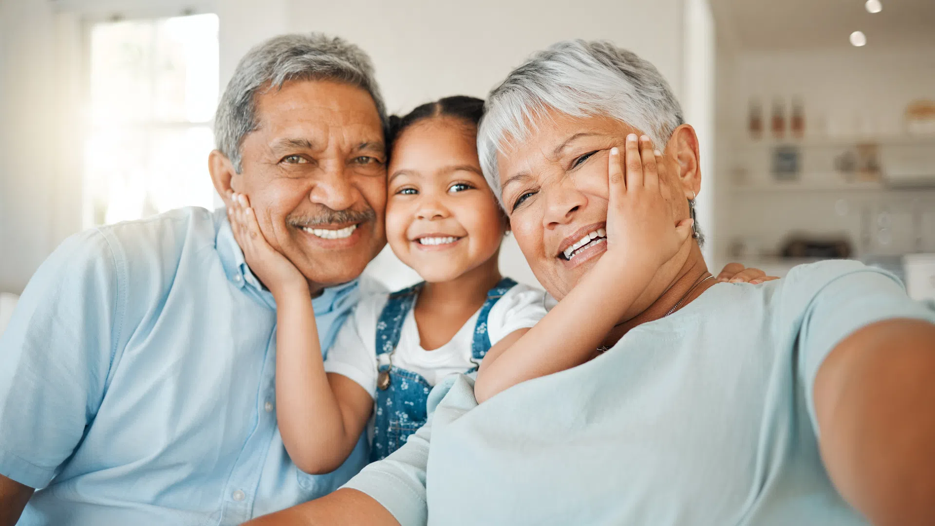 Smiling grandparents and granddaughter taking a happy selfie together at home in a bright room.