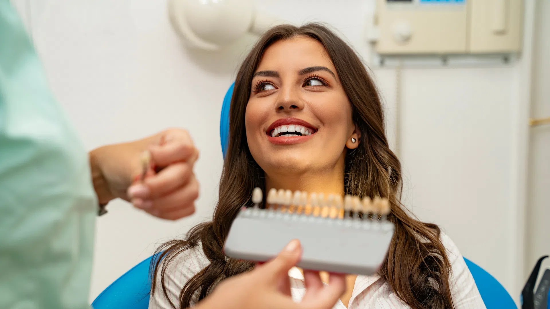 Young woman smiling while dentist shows a dental shade guide for teeth color matching in clinic.