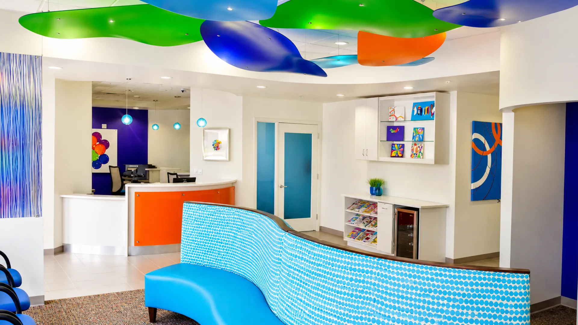 Modern dental office reception with colorful ceiling panels, blue seating, and contemporary artwork on walls.