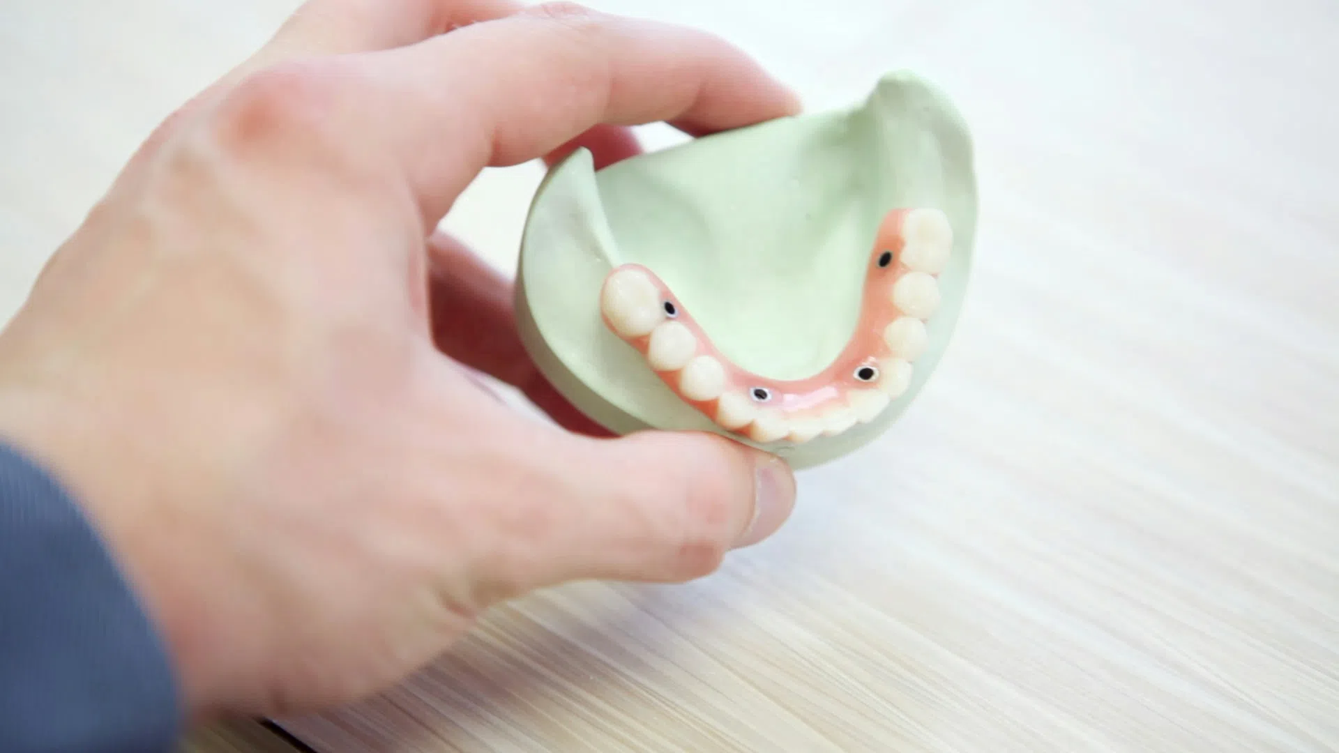 Hand holding a dental mold with artificial lower teeth and implant components on a light table.