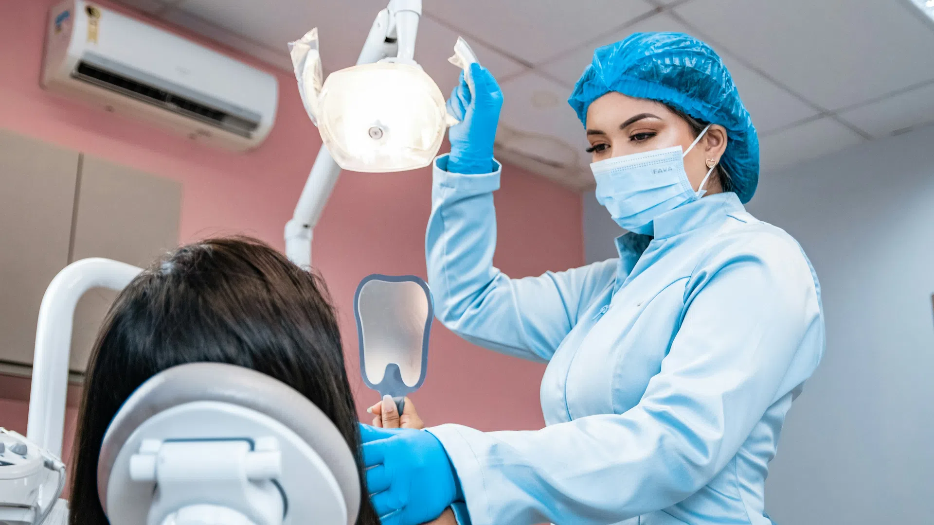 Dentist wearing protective gear adjusts dental light while patient holds mirror in dental office.