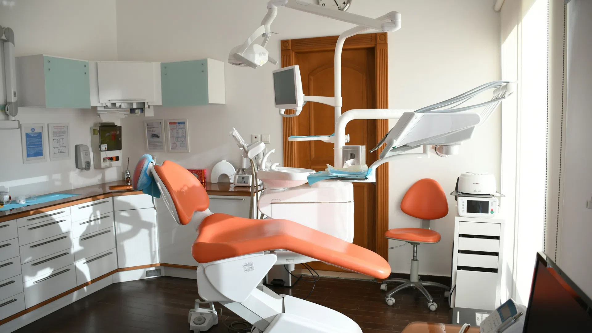 Bright dental clinic room with orange dental chair, equipment, white cabinets, and wooden floor under natural light.