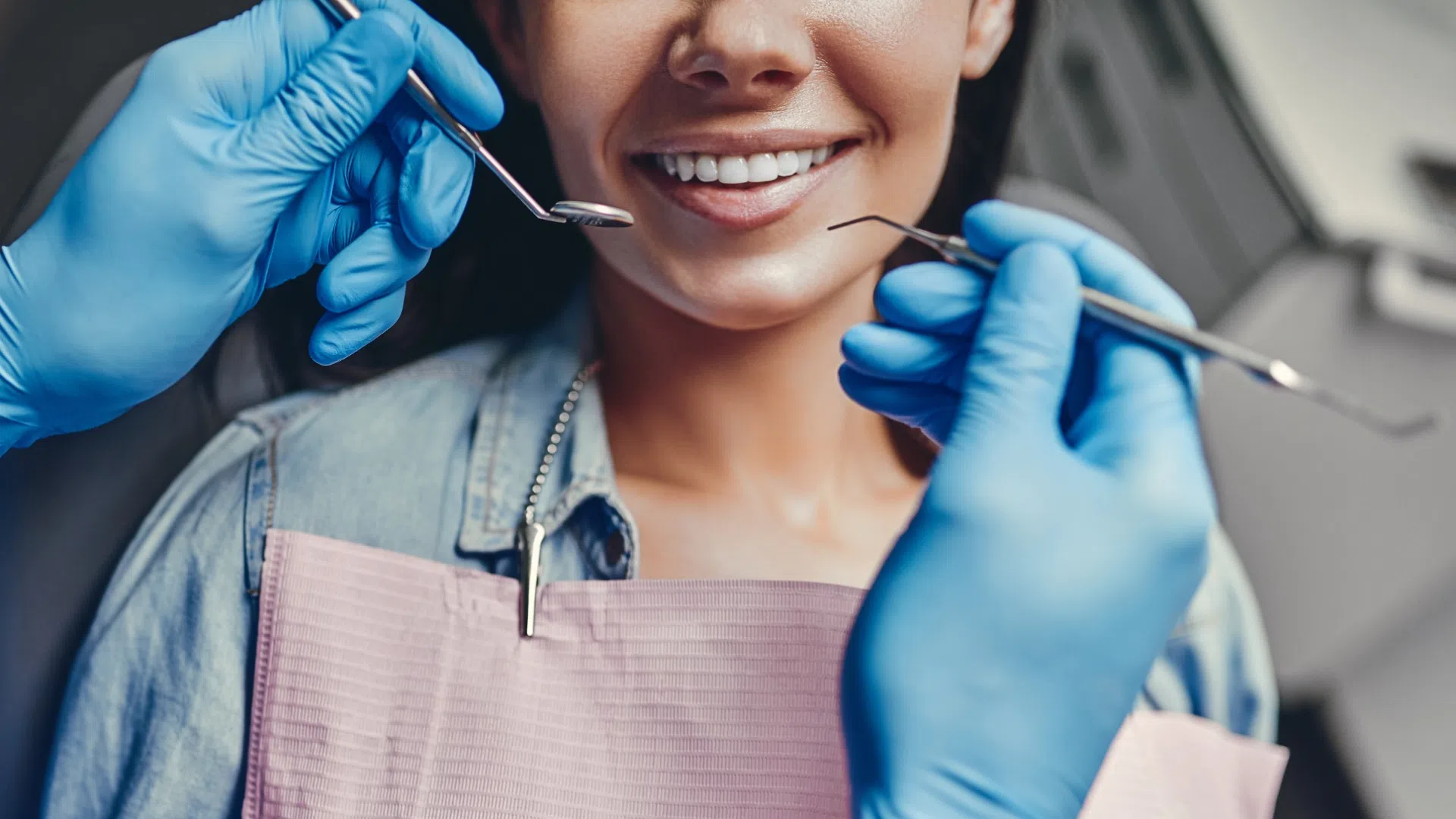 Dentist wearing blue gloves examining a smiling woman's teeth with dental tools at clinic.