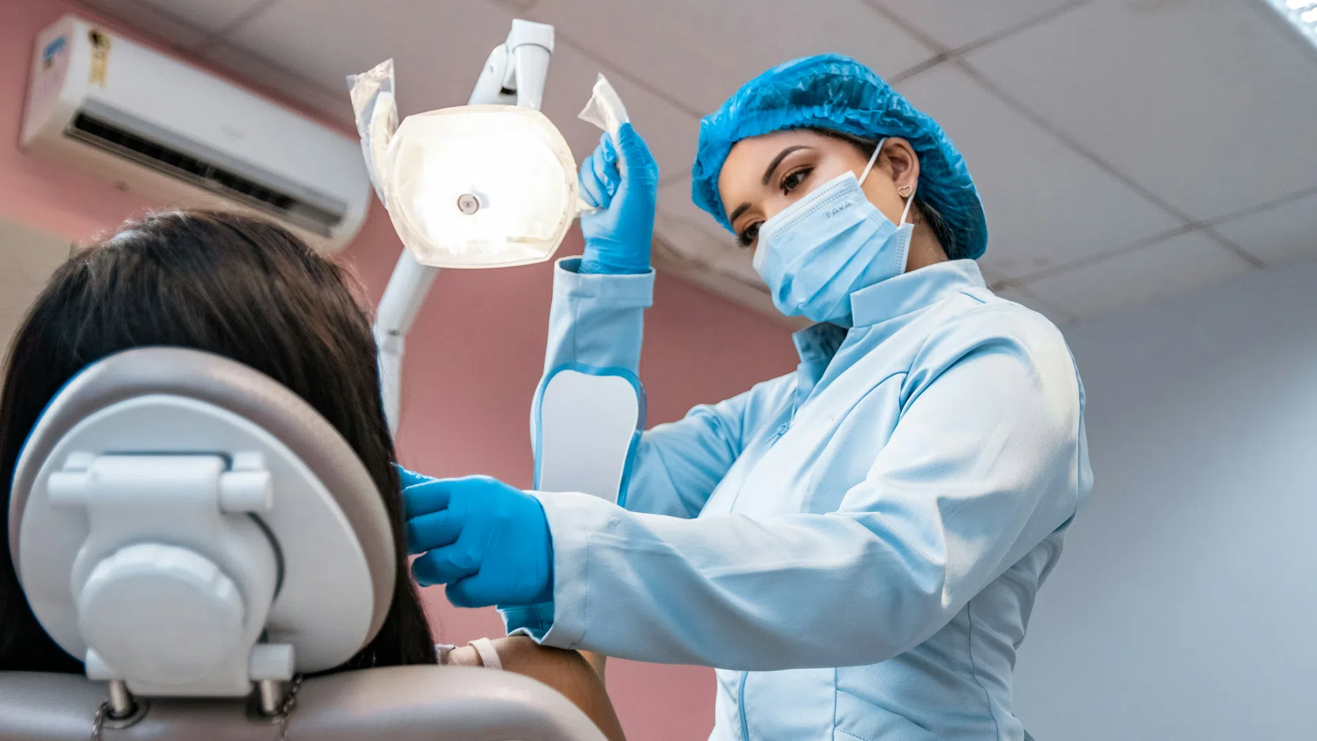Female dentist wearing mask and gloves adjusting overhead light while examining a patient in dental clinic.