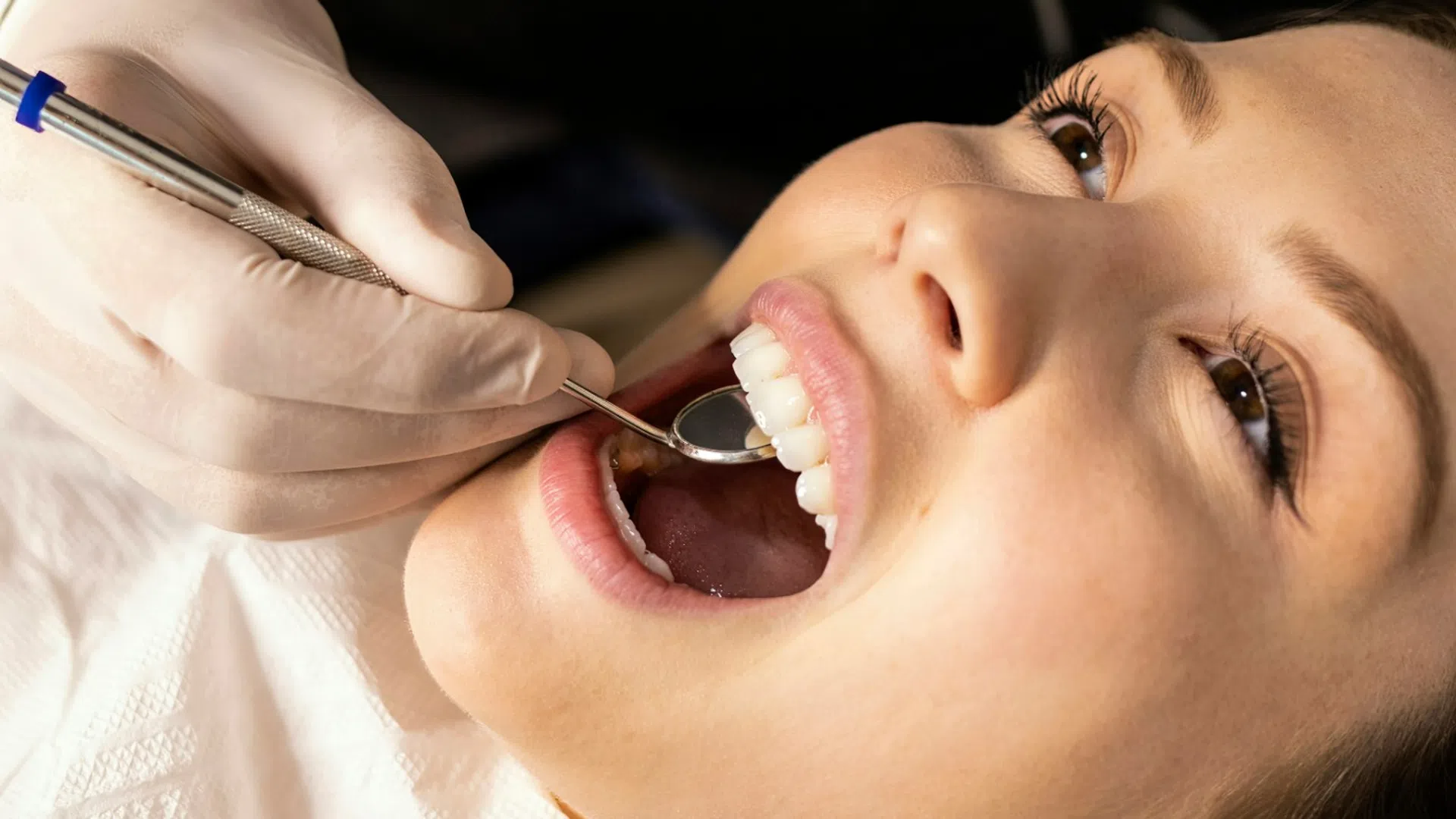 Dentist in gloves examining young woman's teeth with dental mirror during checkup session
