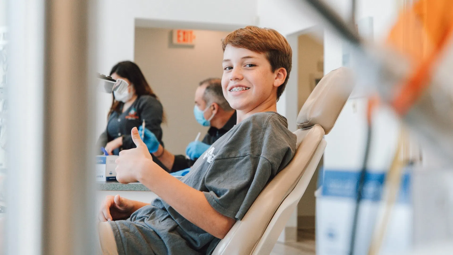 Smiling boy sitting in a dental chair giving thumbs up during a clinic visit with masked staff in background.
