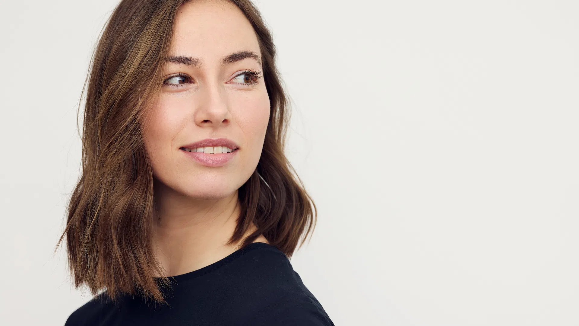 Young woman with shoulder-length brown hair wearing a black shirt looking to the side against a white background