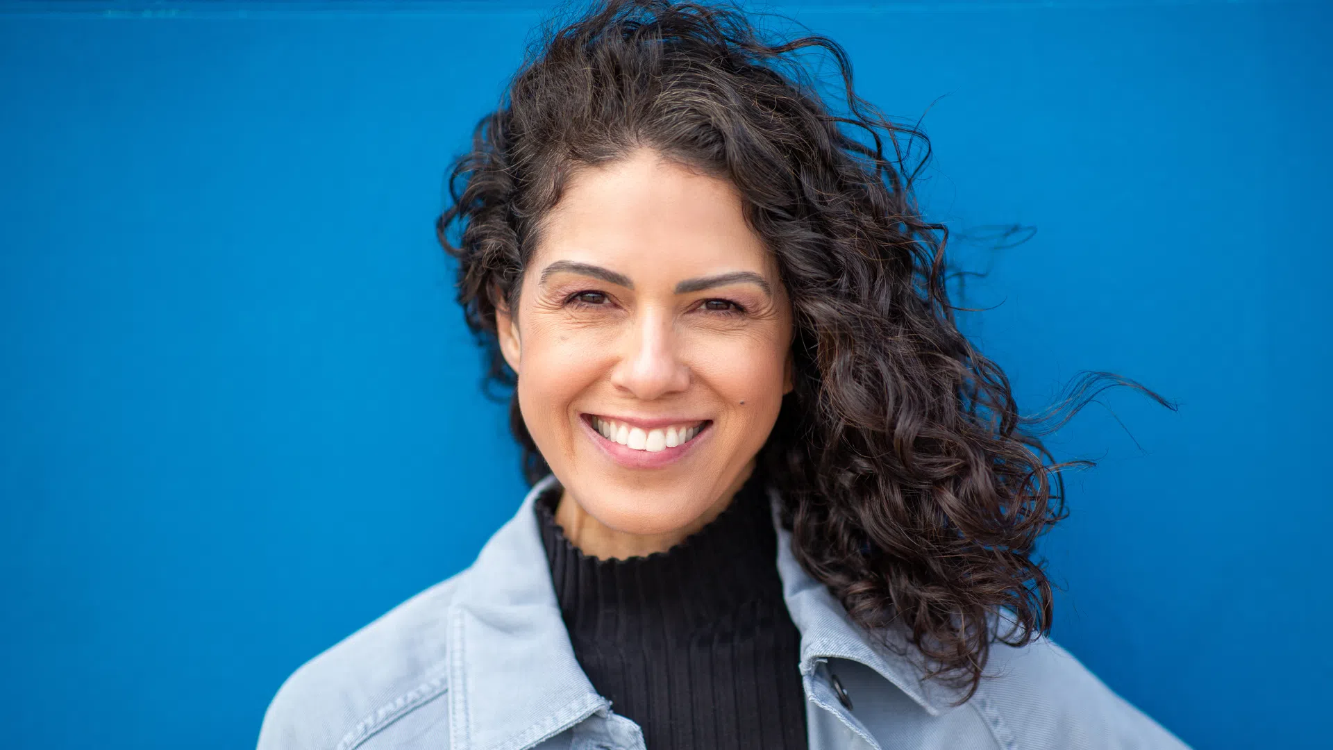 Smiling woman with curly dark hair wearing a light jacket and black top against a blue background