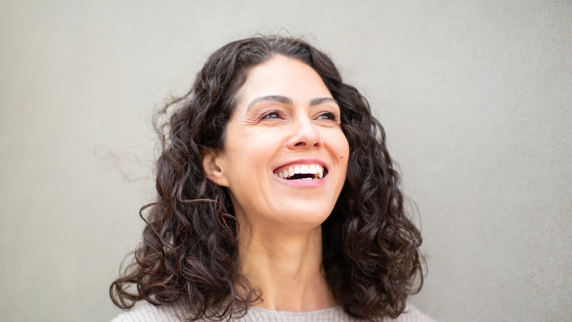 Happy woman with curly dark hair smiling against a plain beige wall background.