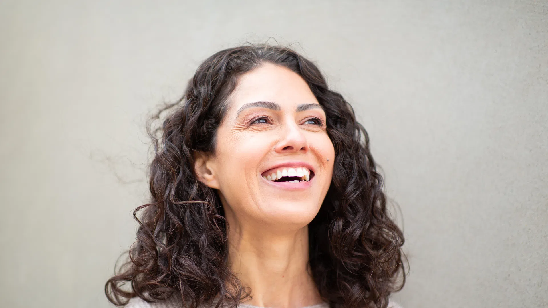Happy woman with curly dark hair smiling against a plain beige wall background.