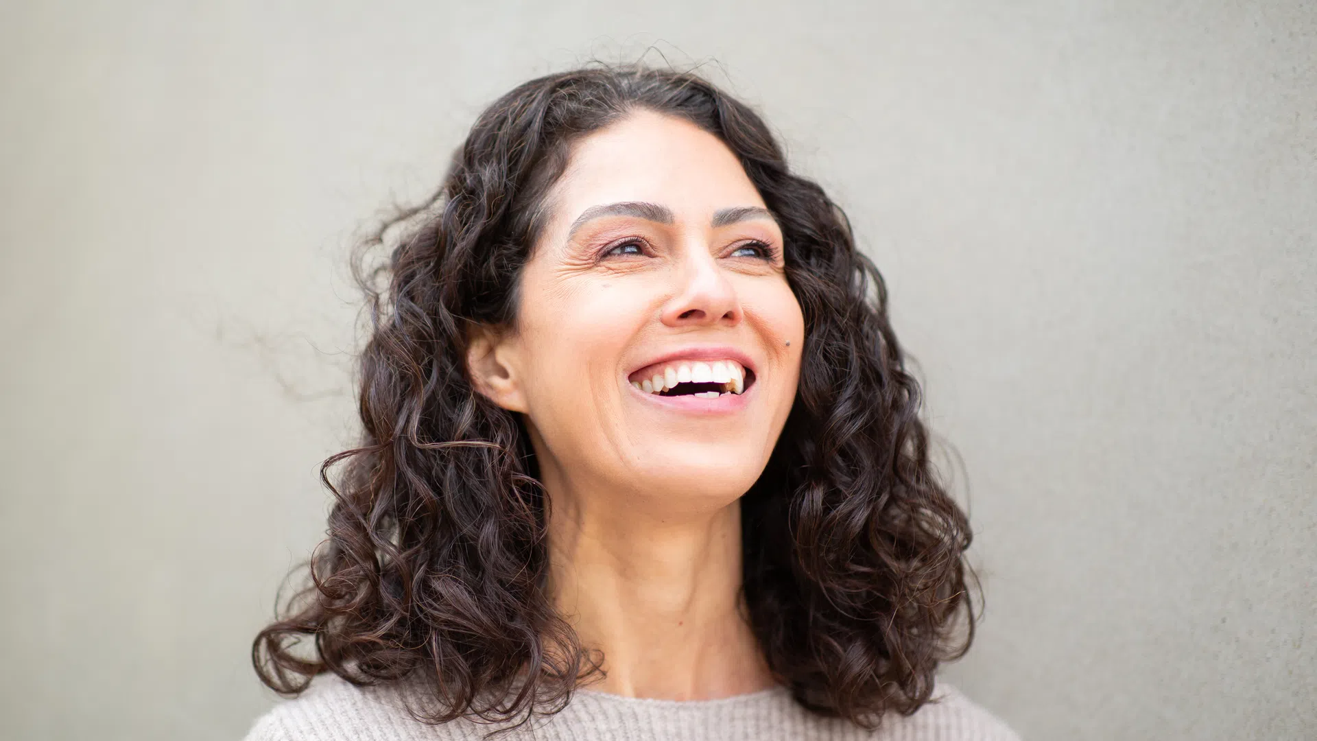 Happy woman with curly dark hair smiling against a plain beige wall background.