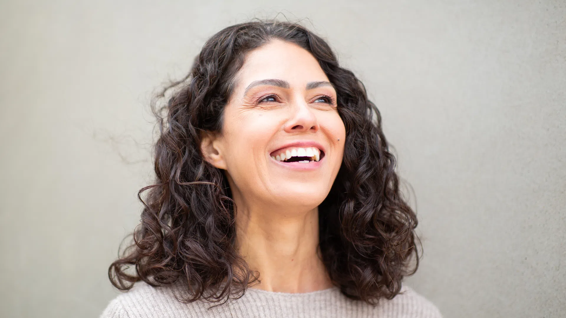 Happy woman with curly dark hair smiling against a plain beige wall background.