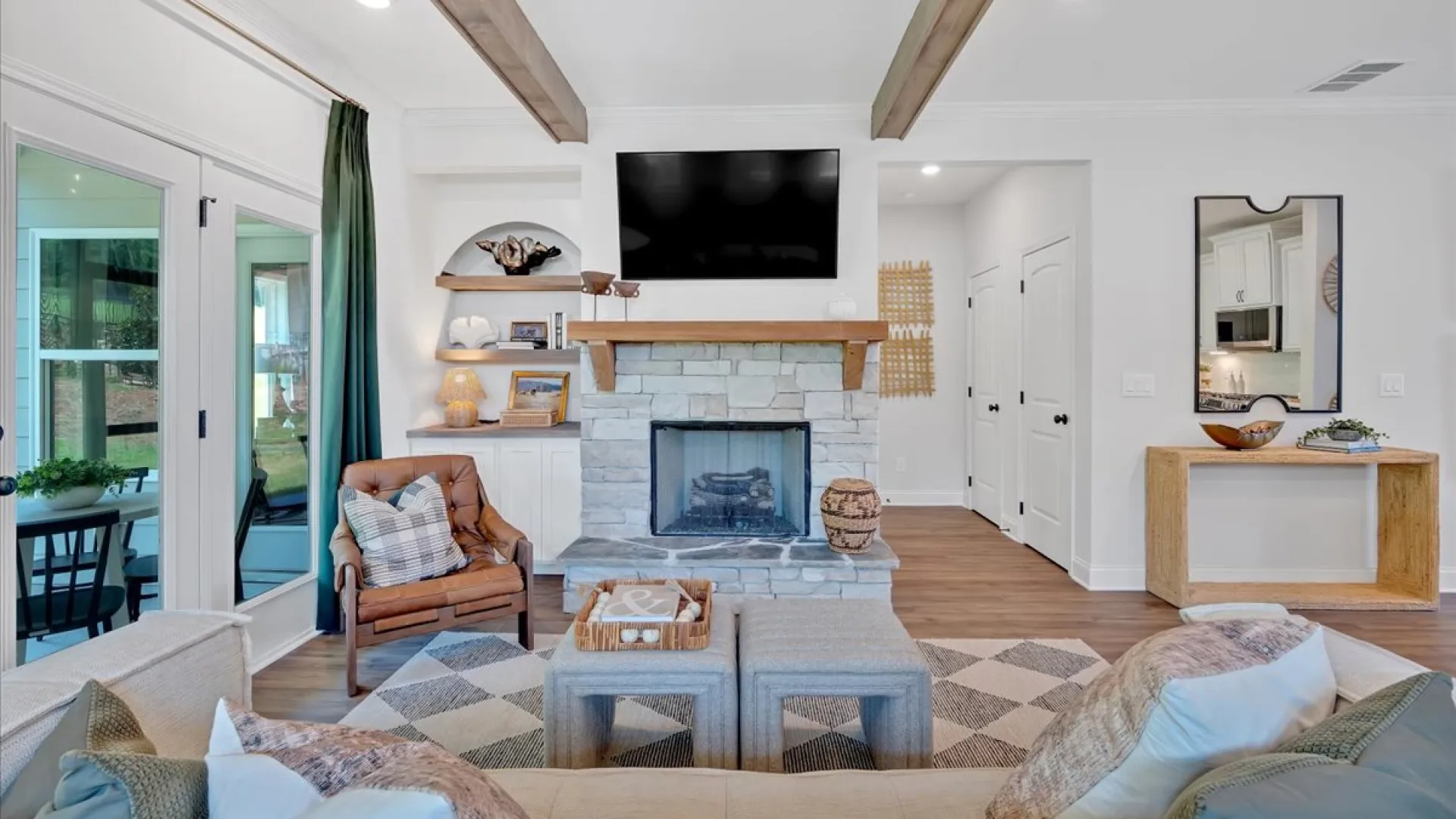 Bright modern living room with stone fireplace, mounted TV, cozy seating, wooden beams, and large windows.