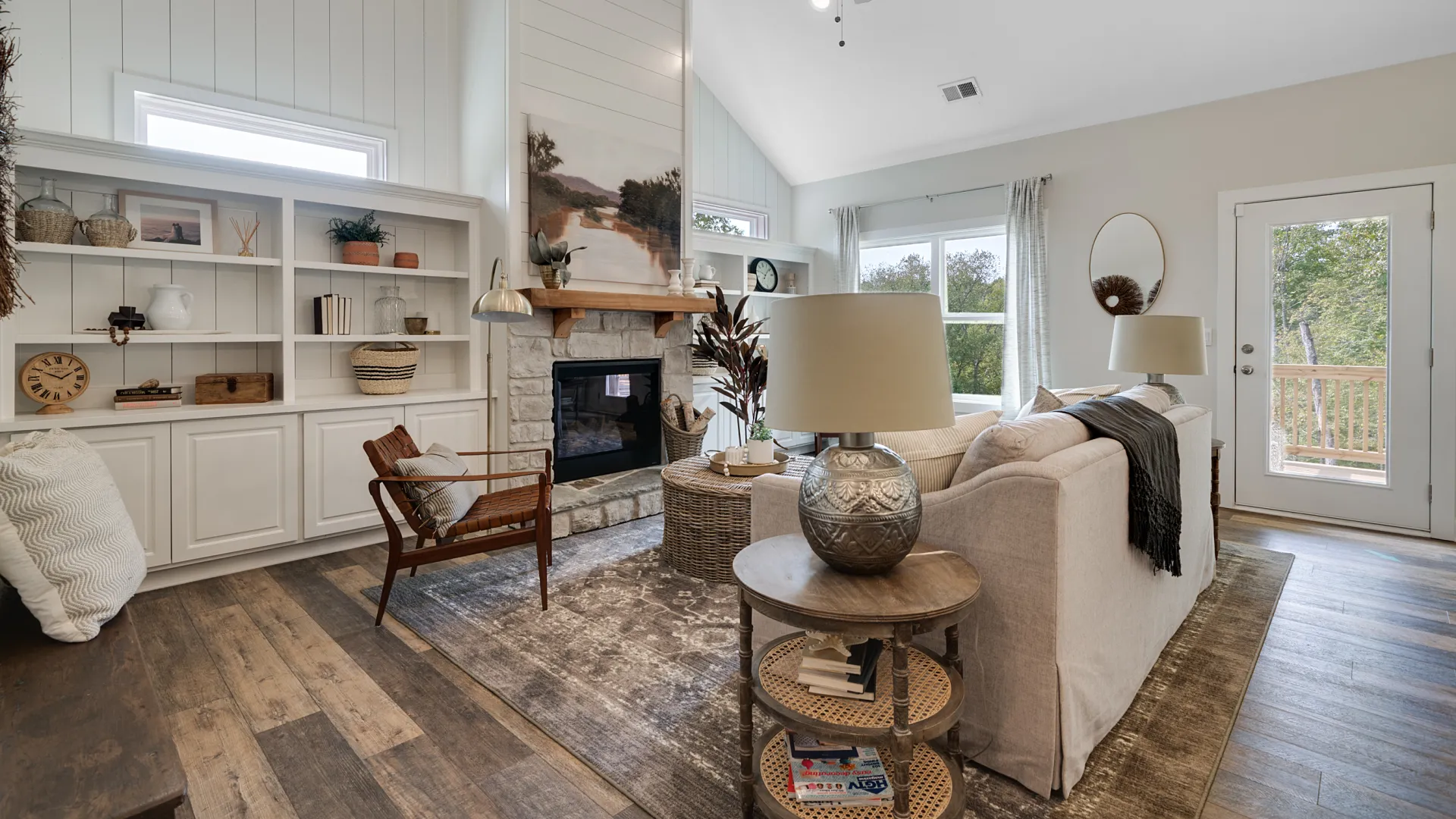 Bright living room with vaulted ceiling, stone fireplace, wooden furniture, and large windows overlooking greenery.