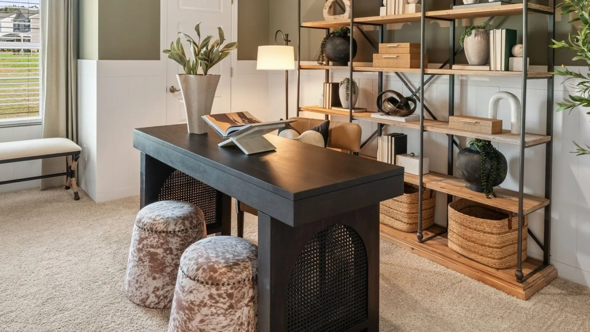 Modern home office with black desk, open shelving, beige carpet, bench, and potted plants under natural light.
