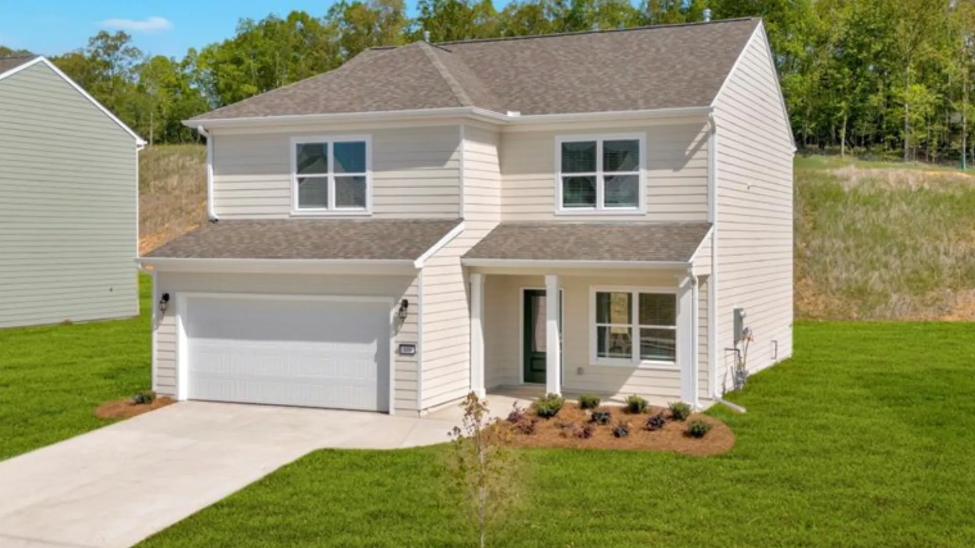Two-story beige house with white garage door, green lawn, and small landscaped bushes under a blue sky.