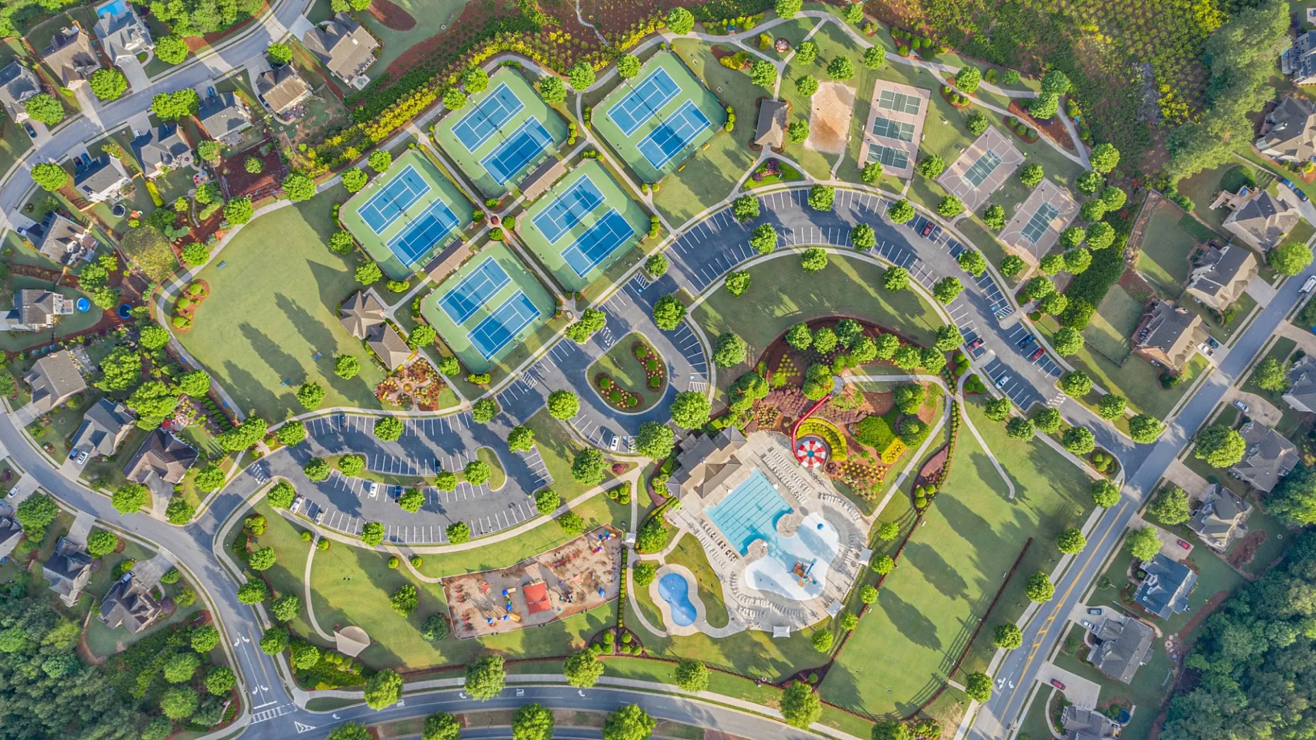 Aerial view of a suburban neighborhood featuring multiple tennis courts, a pool, playground, and curved roads.