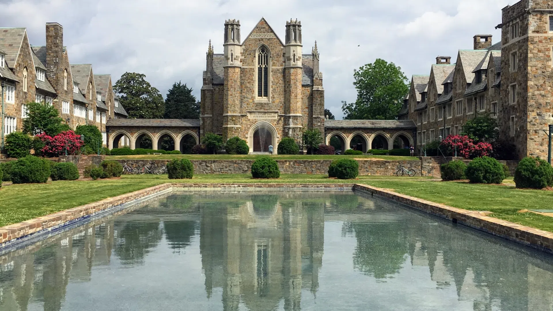 A historic building reflected in a serene pond surrounded by lush greenery and vibrant flowers.