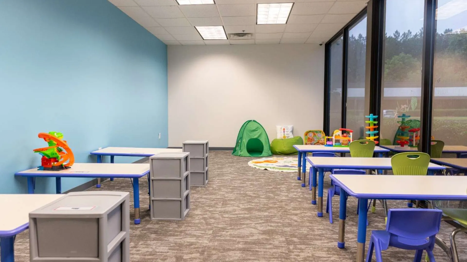 Bright classroom with blue and green chairs, white desks, toys, and a green play tent near large windows.