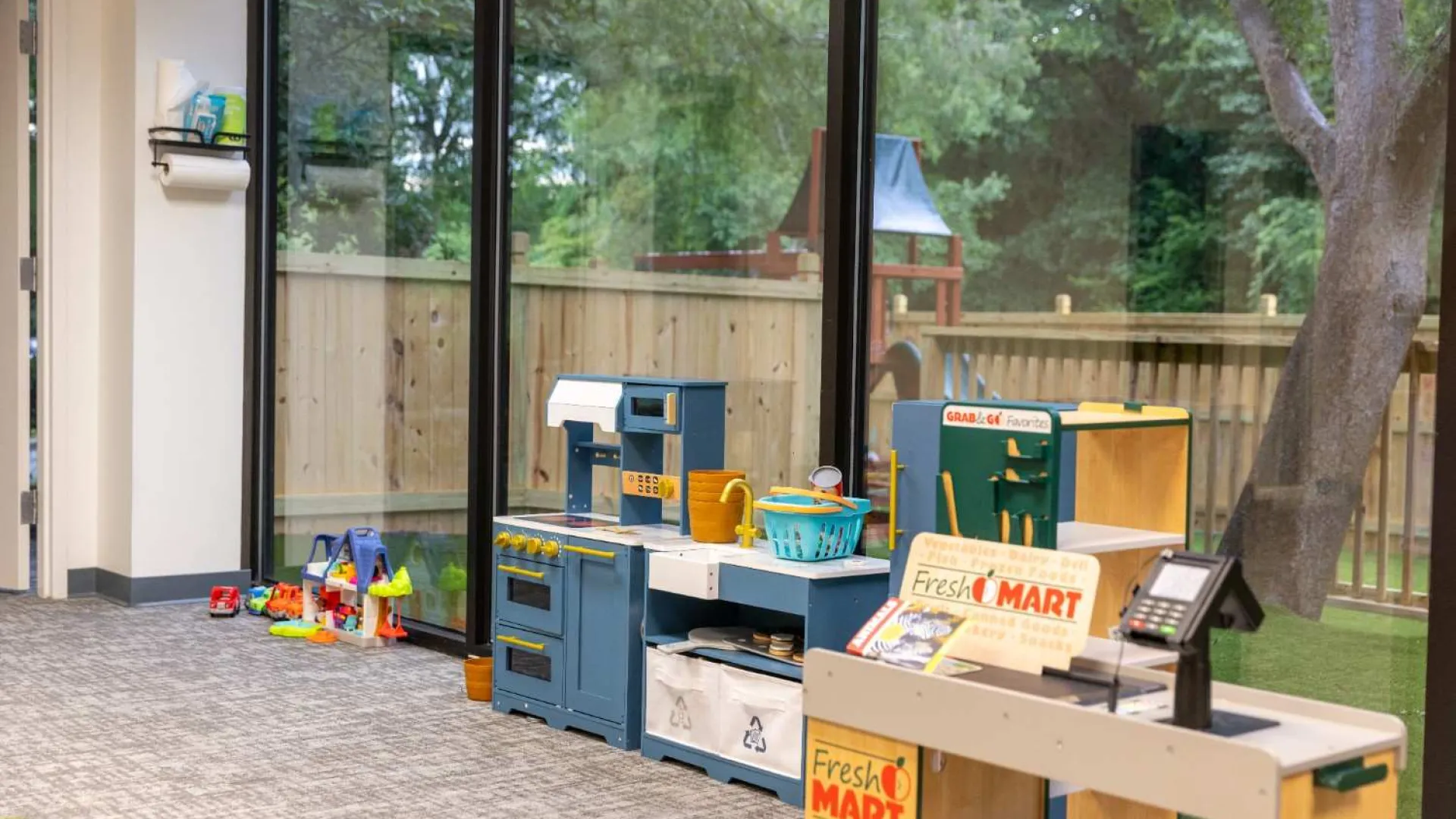 Children's play area with toy kitchen sets and a small grocery store near large windows overlooking a backyard.