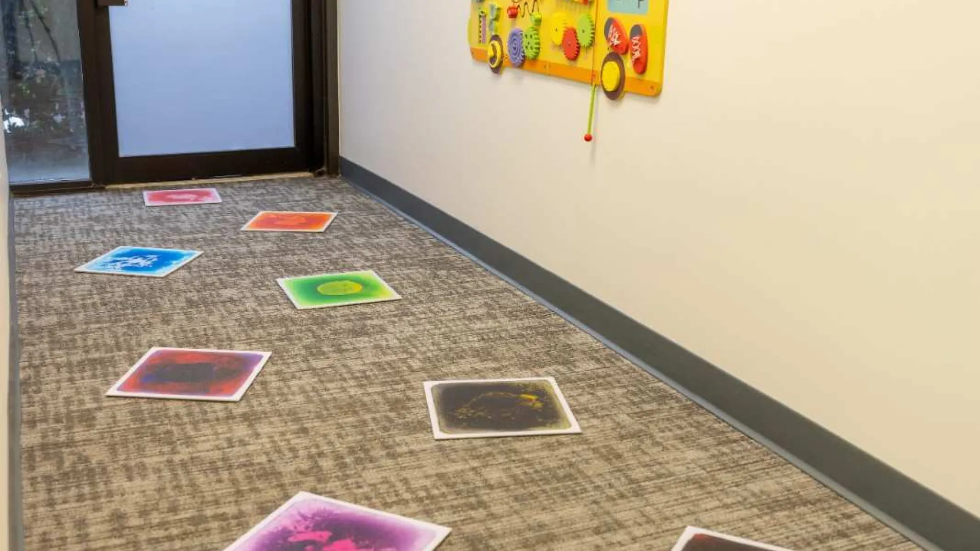 Colorful sensory activity wall and vibrant stepping stones on carpeted hallway floor near glass door
