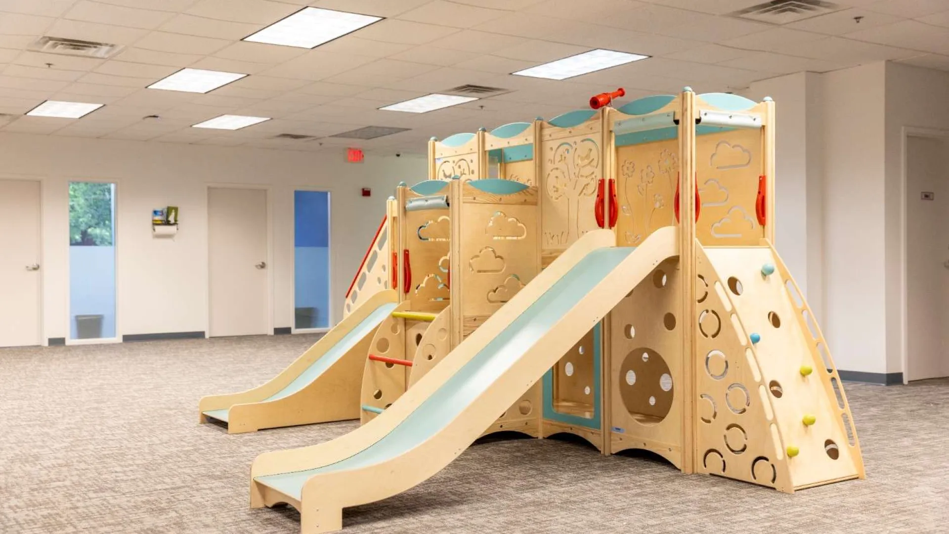 Indoor wooden play structure with slides and climbing elements in a bright modern room.