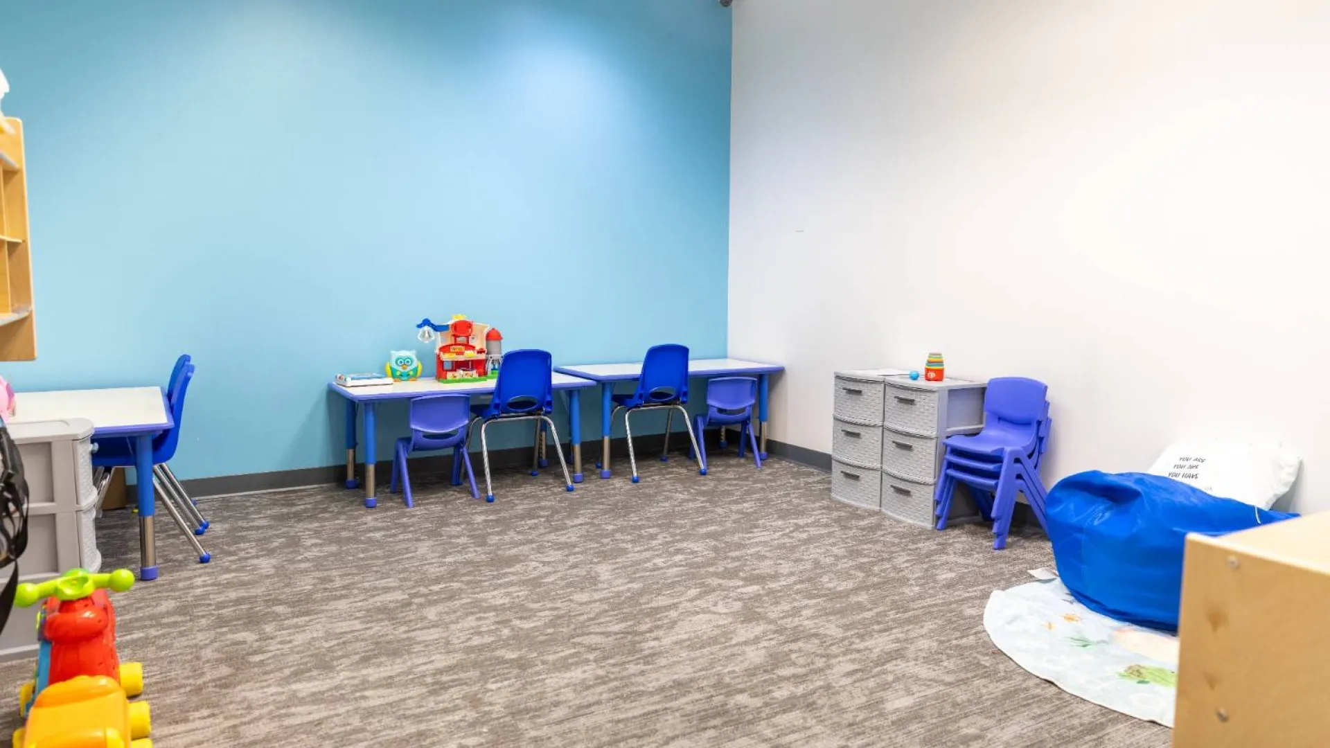 Bright and organized children's play and learning room with small tables, blue chairs, toys, and bean bag seating.