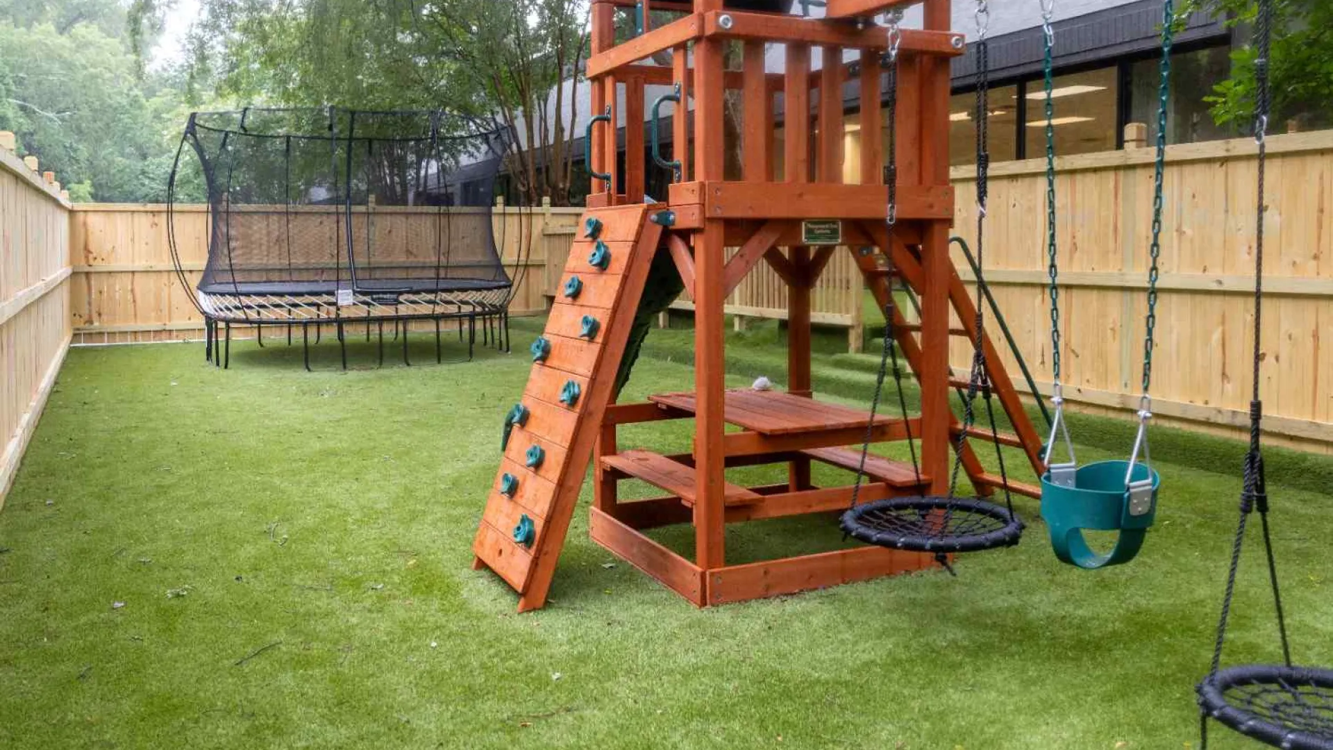 Backyard playground set with swings, climbing wall, picnic table, and trampoline on green lawn surrounded by wooden fence