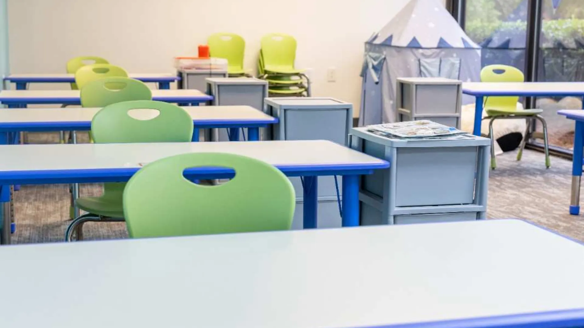 Empty classroom with green chairs, blue-trimmed desks, gray storage bins, and a play tent by large windows.