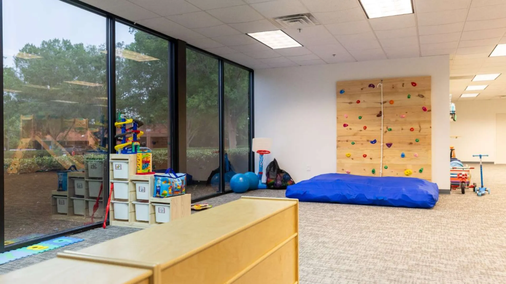 Bright indoor playroom with climbing wall, large windows, toys, and padded floor mat for children's activities.