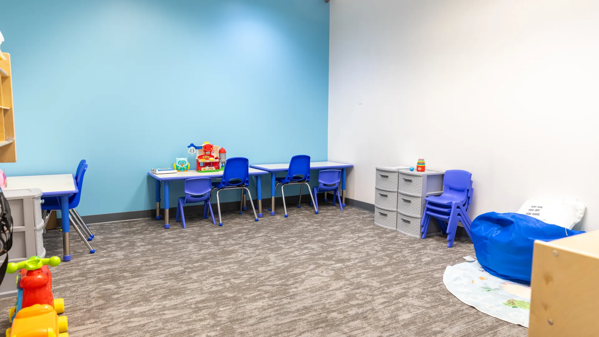 Bright and clean children's classroom with small blue chairs, desks, toys, and a bean bag chair on carpeted floor