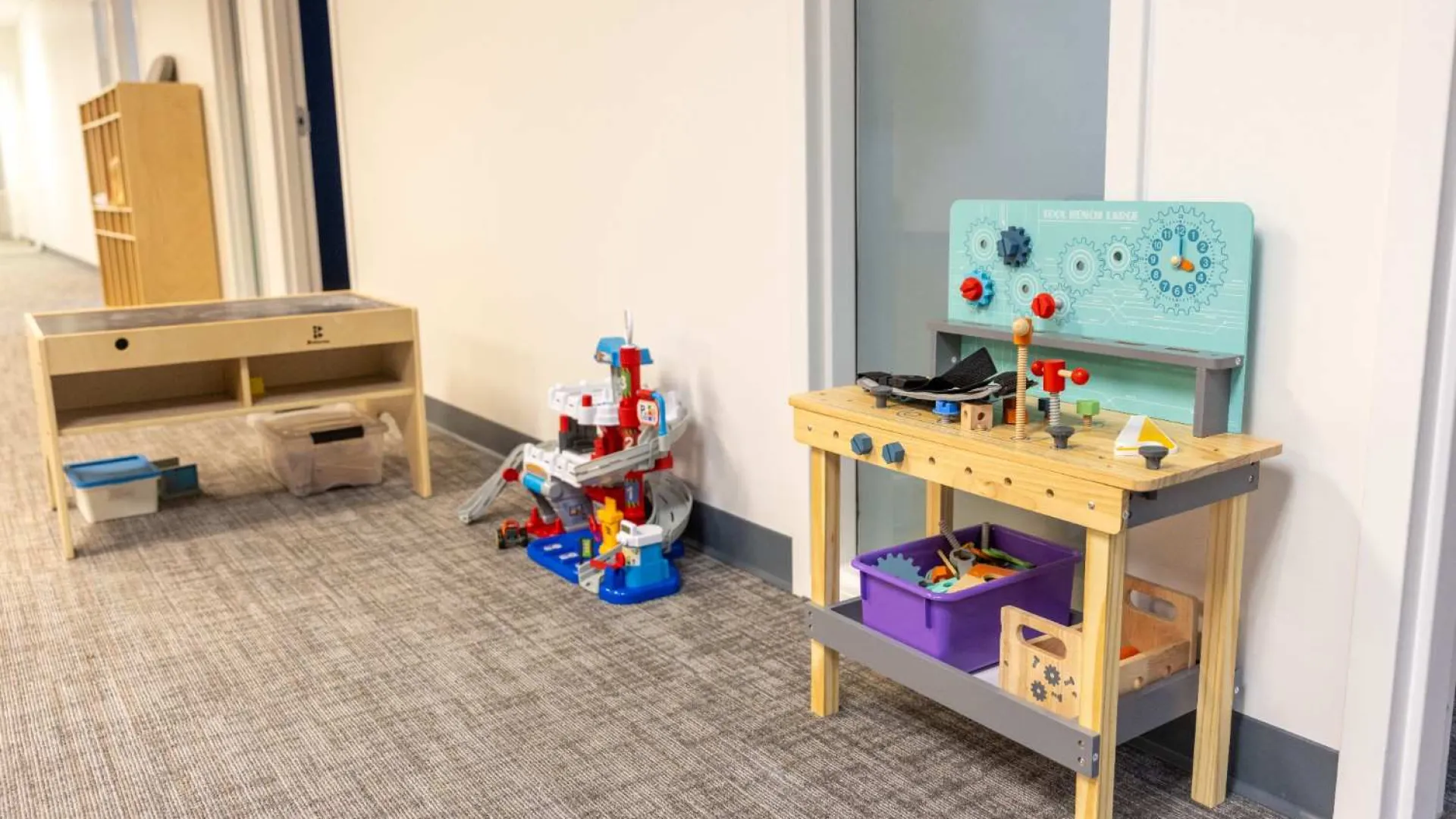 Children's play area with wooden activity table, toy tools, and colorful slide set against hallway wall.