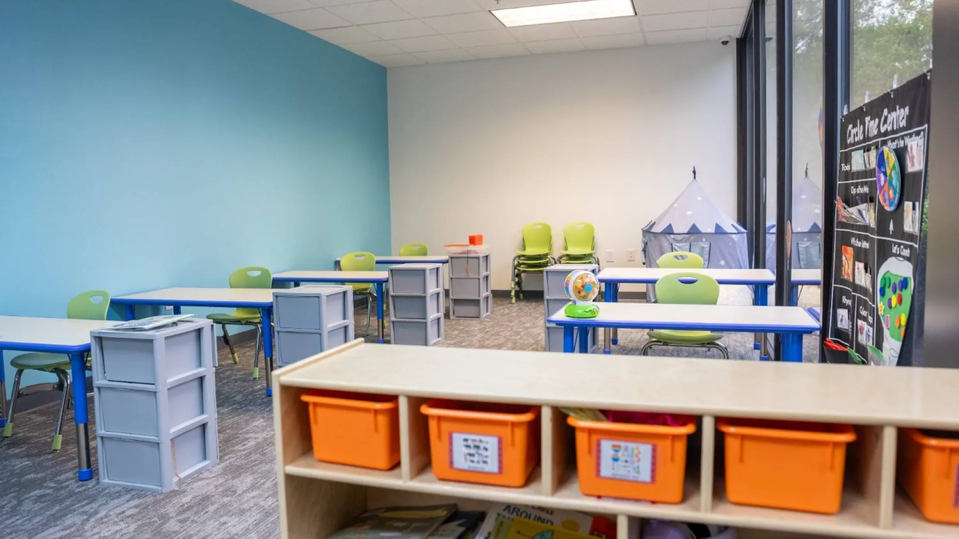 Bright classroom with blue walls, green chairs, small desks, orange storage bins, and a play tent by the window.
