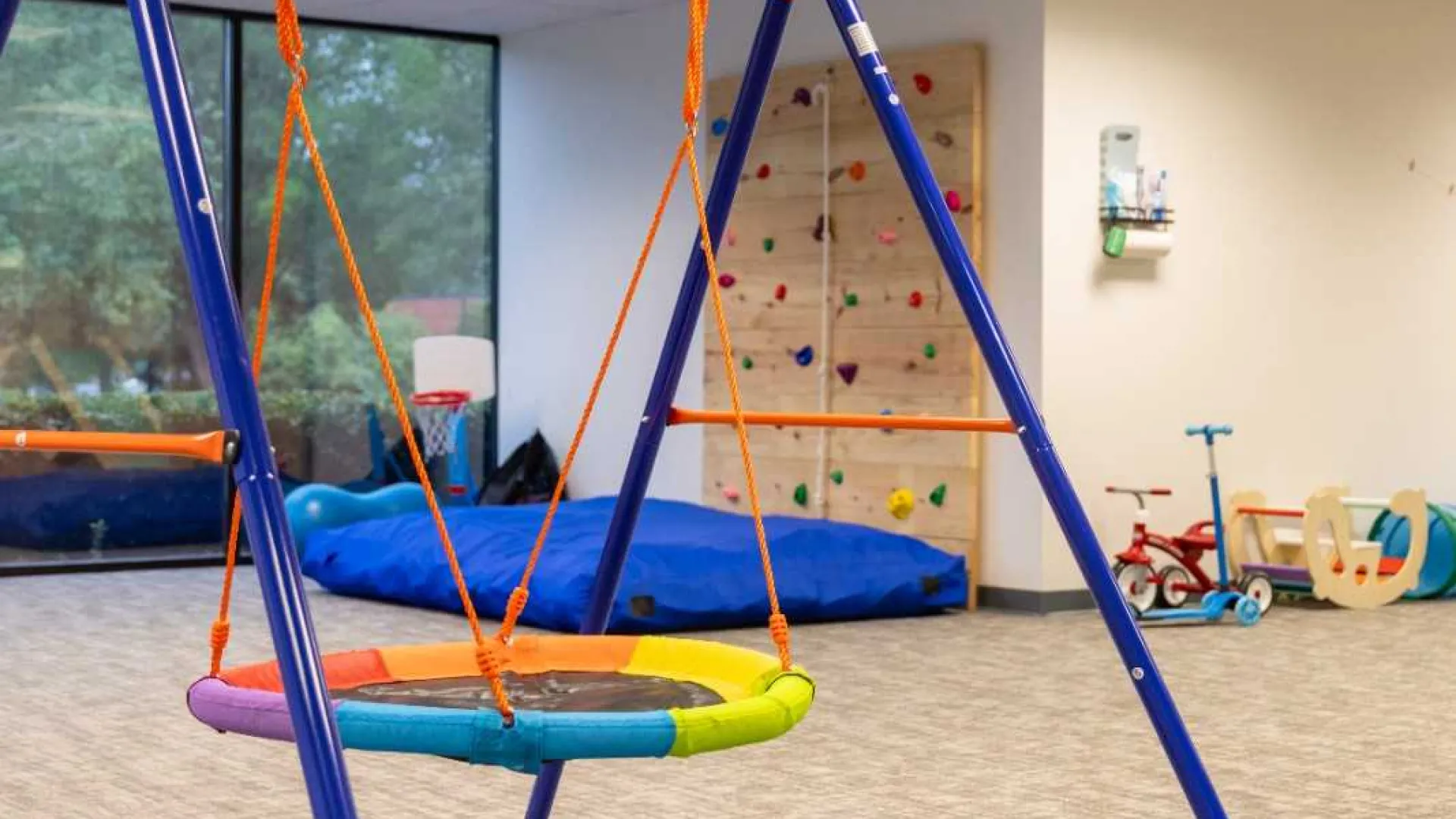 Indoor play area with a colorful round swing, climbing wall, and children's toys in a bright room.