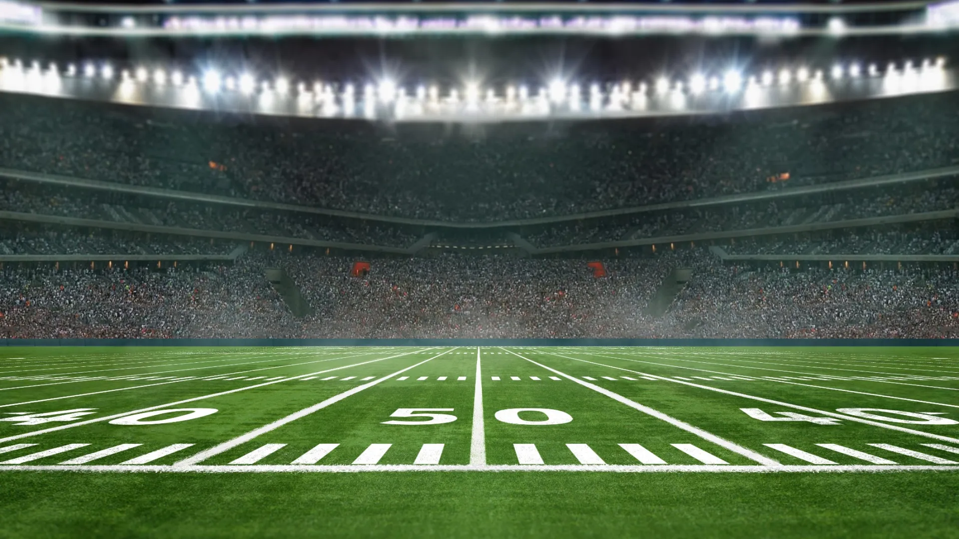 View of a brightly lit American football stadium with a large crowd and marked green field at the 50-yard line.