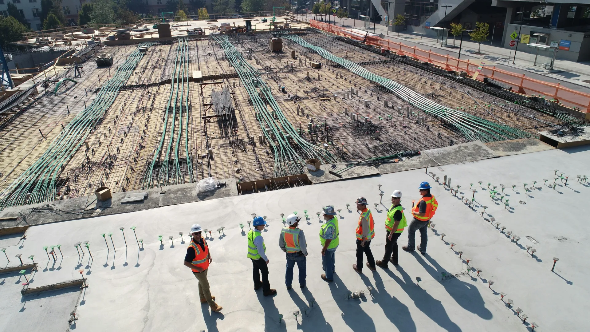 Construction workers in safety gear inspecting large concrete foundation with rebar and conduit layout outdoors.