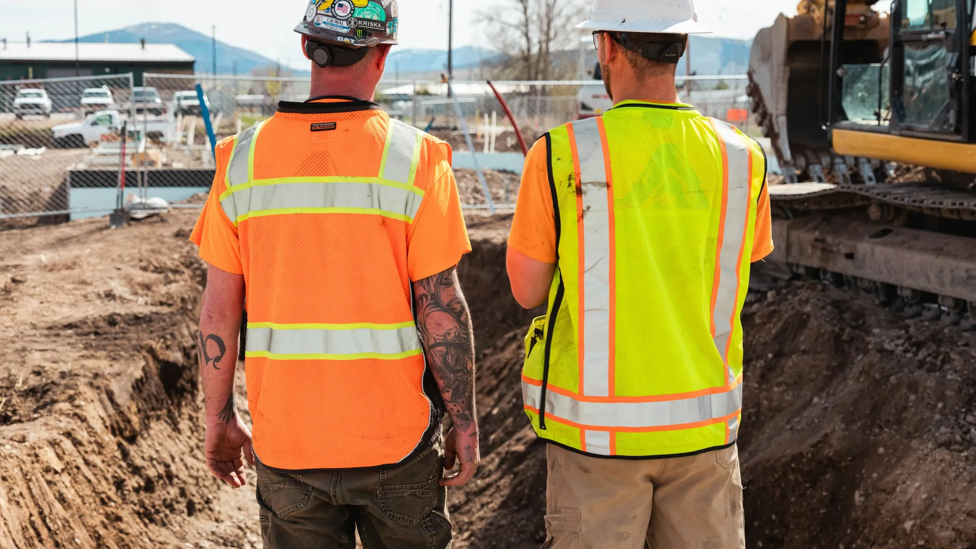 Two construction workers in safety vests and helmets standing by an excavation site with heavy machinery.