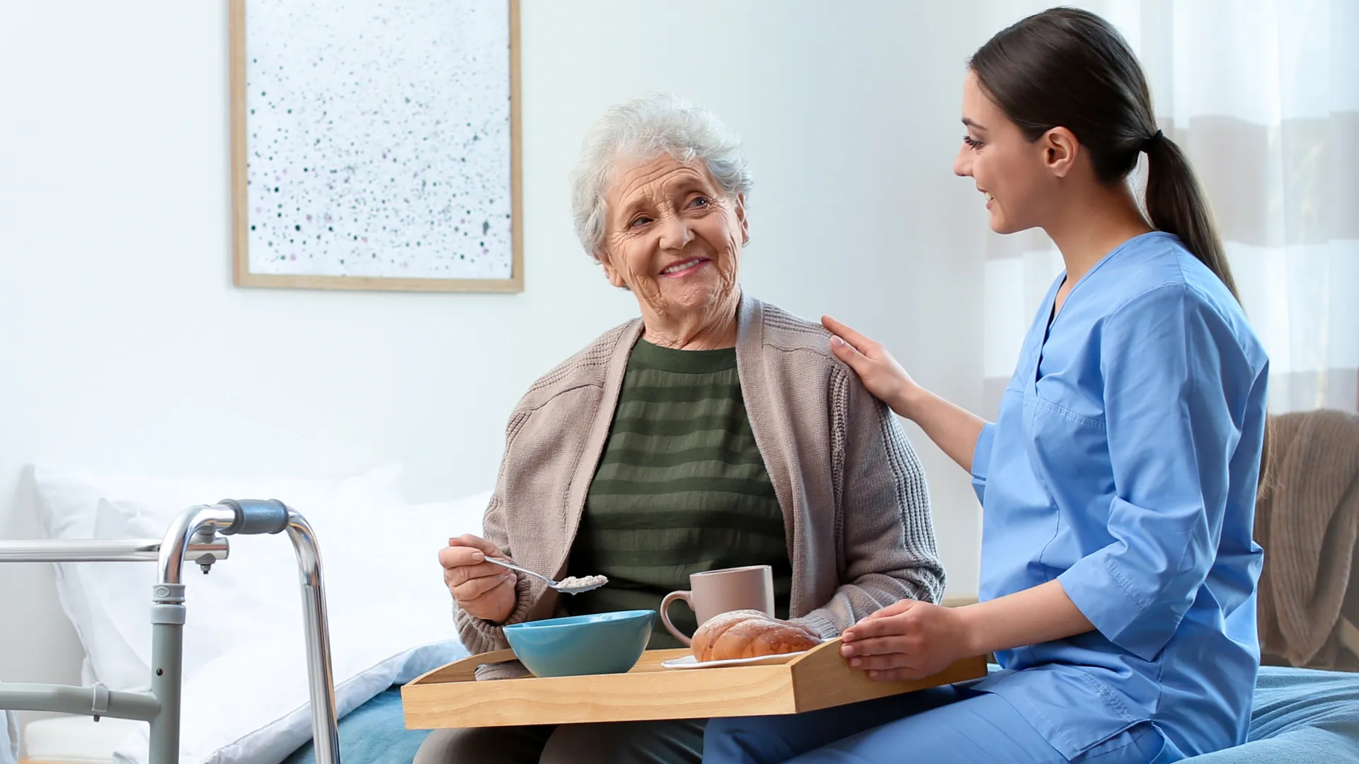 Elderly woman and caregiver sharing a meal and smiling in a cozy living room with walker nearby.