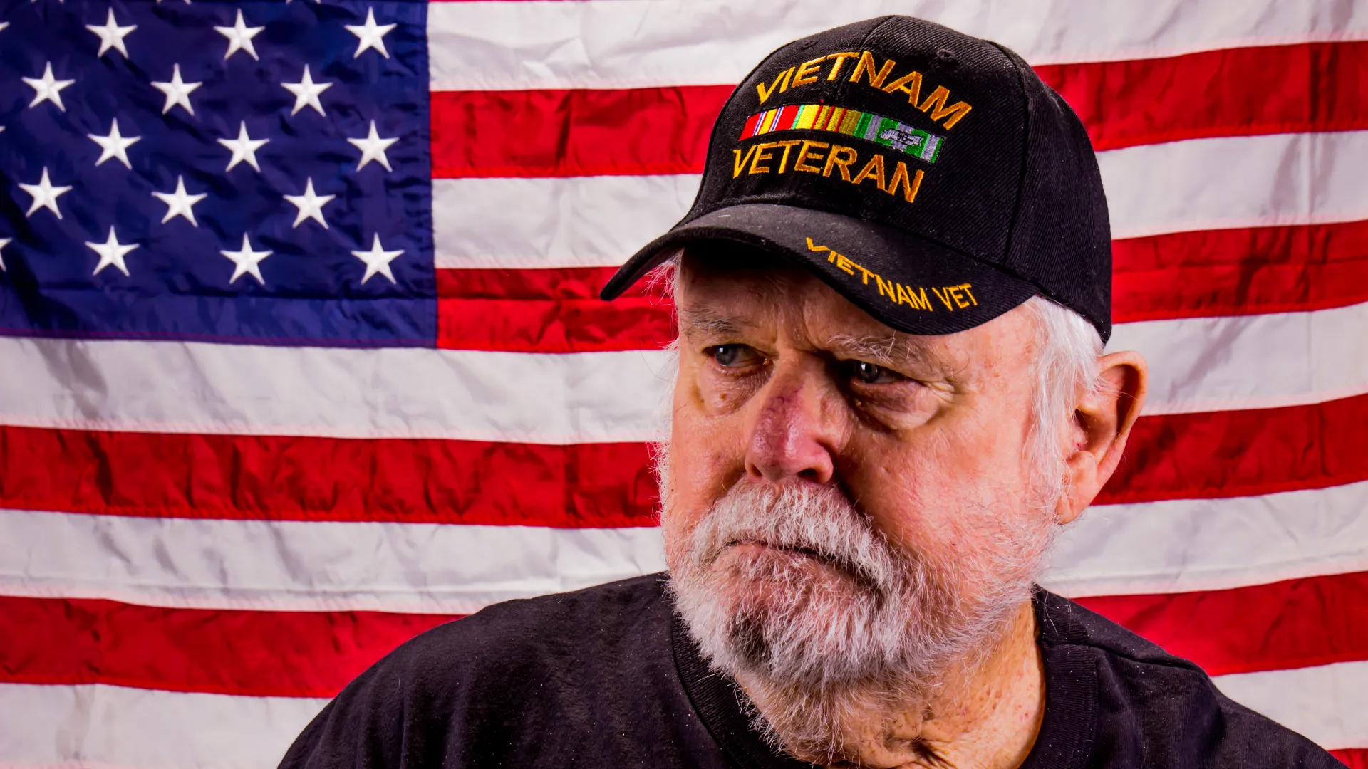 Elderly man wearing a Vietnam Veteran hat in front of a large American flag with a serious expression.
