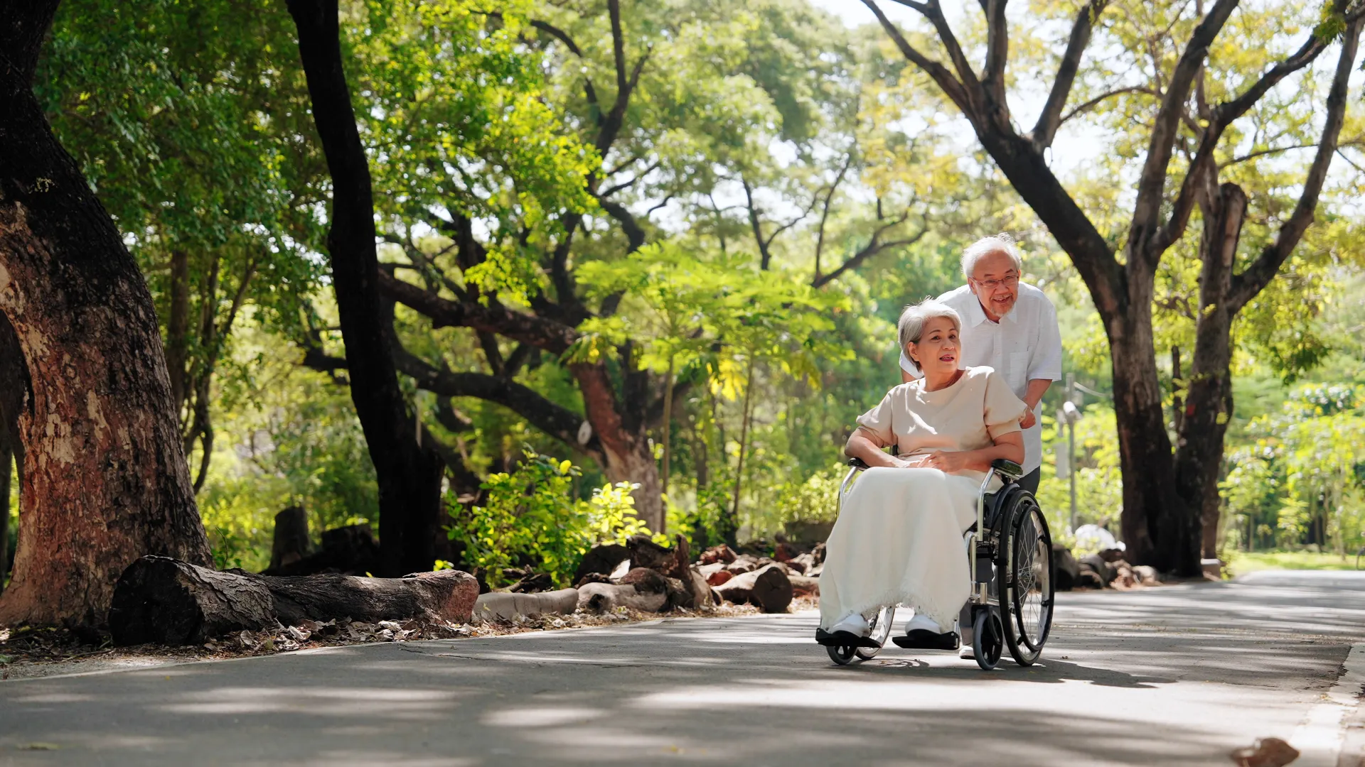 Elderly man pushing a woman in a wheelchair along a sunlit park path surrounded by green trees.