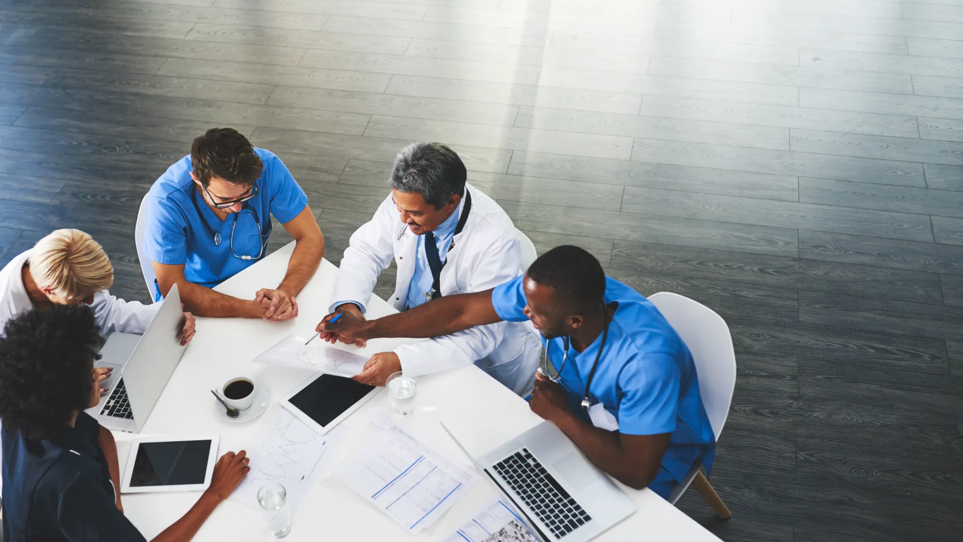 Medical team in blue scrubs and white coat discussing patient charts and using laptops around a white table.