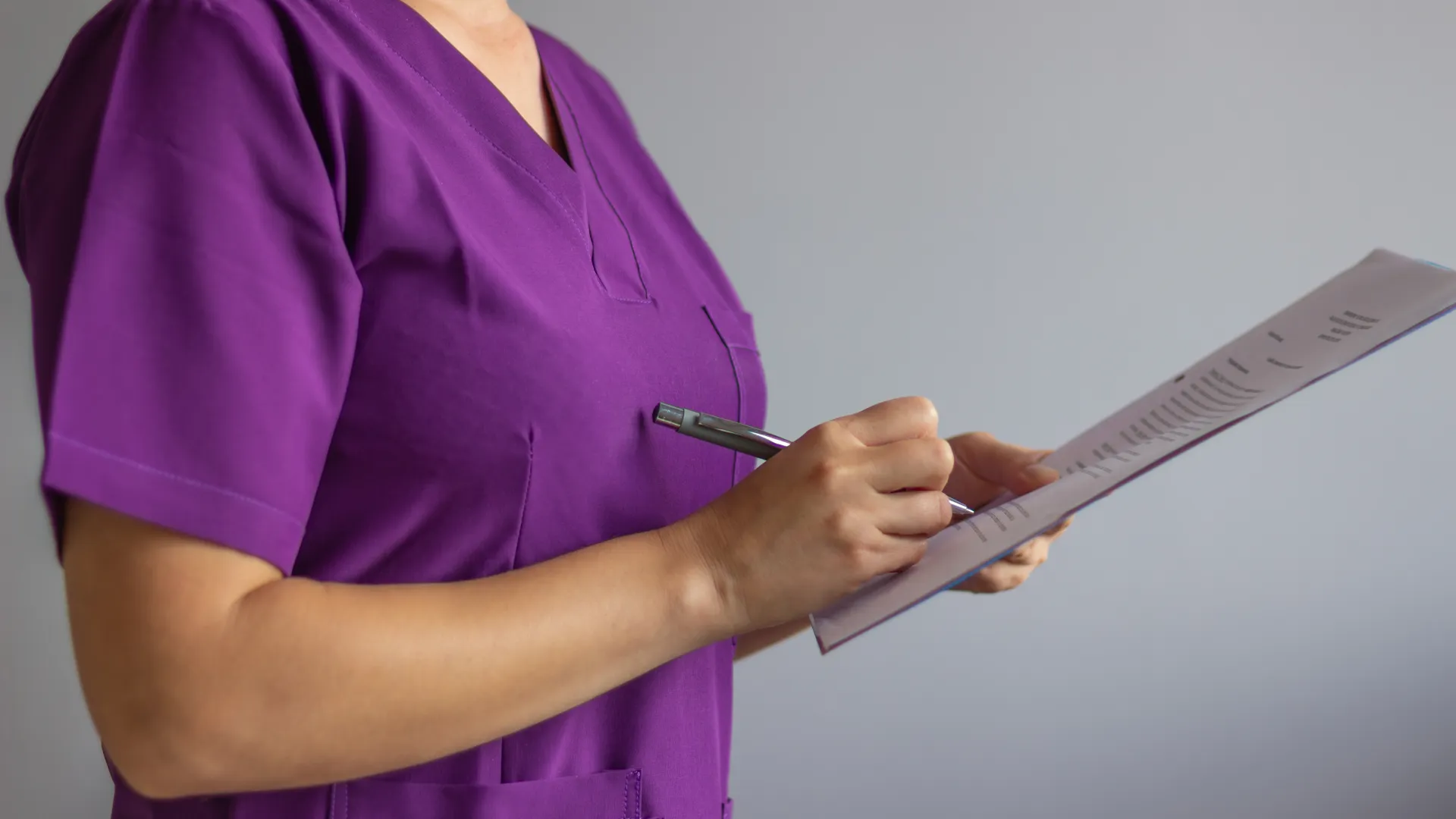 Medical professional in purple scrubs writing on a clipboard against a gray background