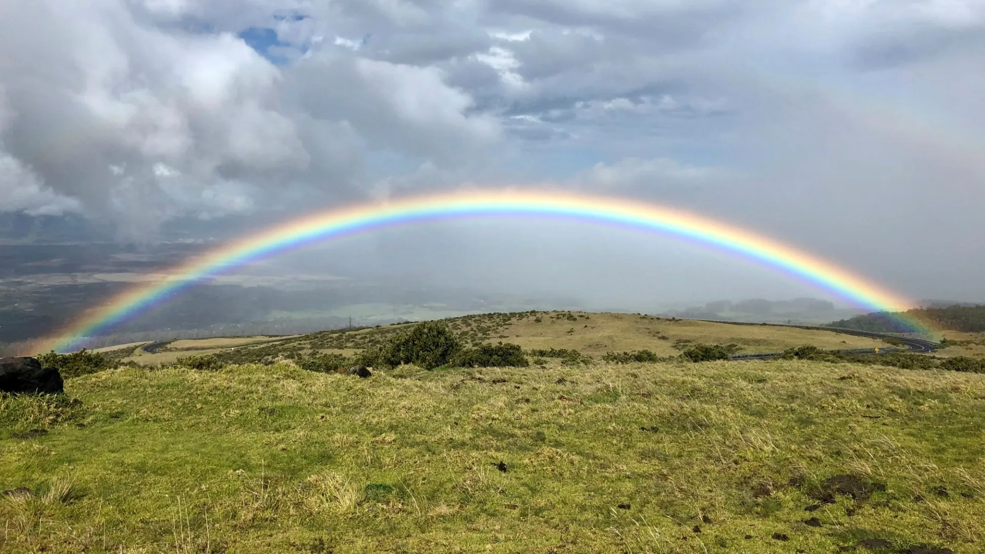 Bright rainbow arching over a green grassy hillside under a cloudy sky with patches of blue.