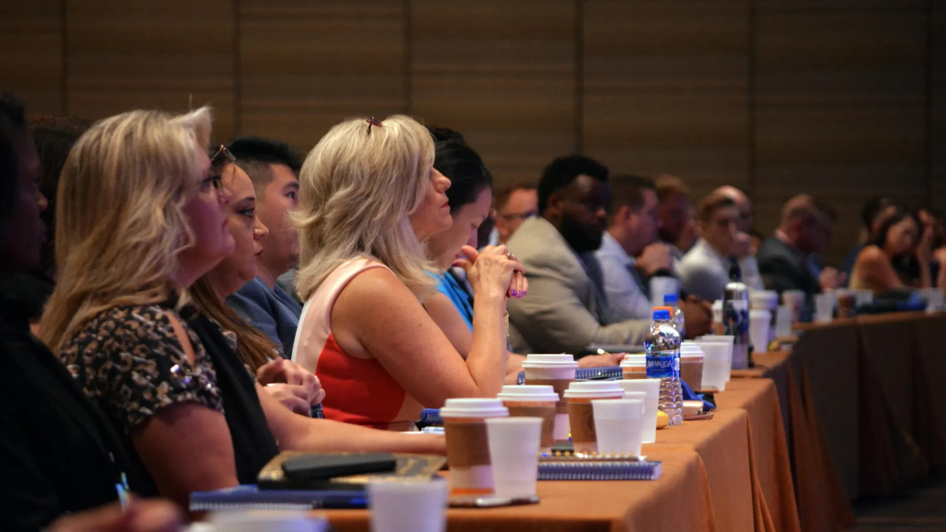 Diverse group of professionals attentively listening at a conference with notebooks and coffee cups on the table.