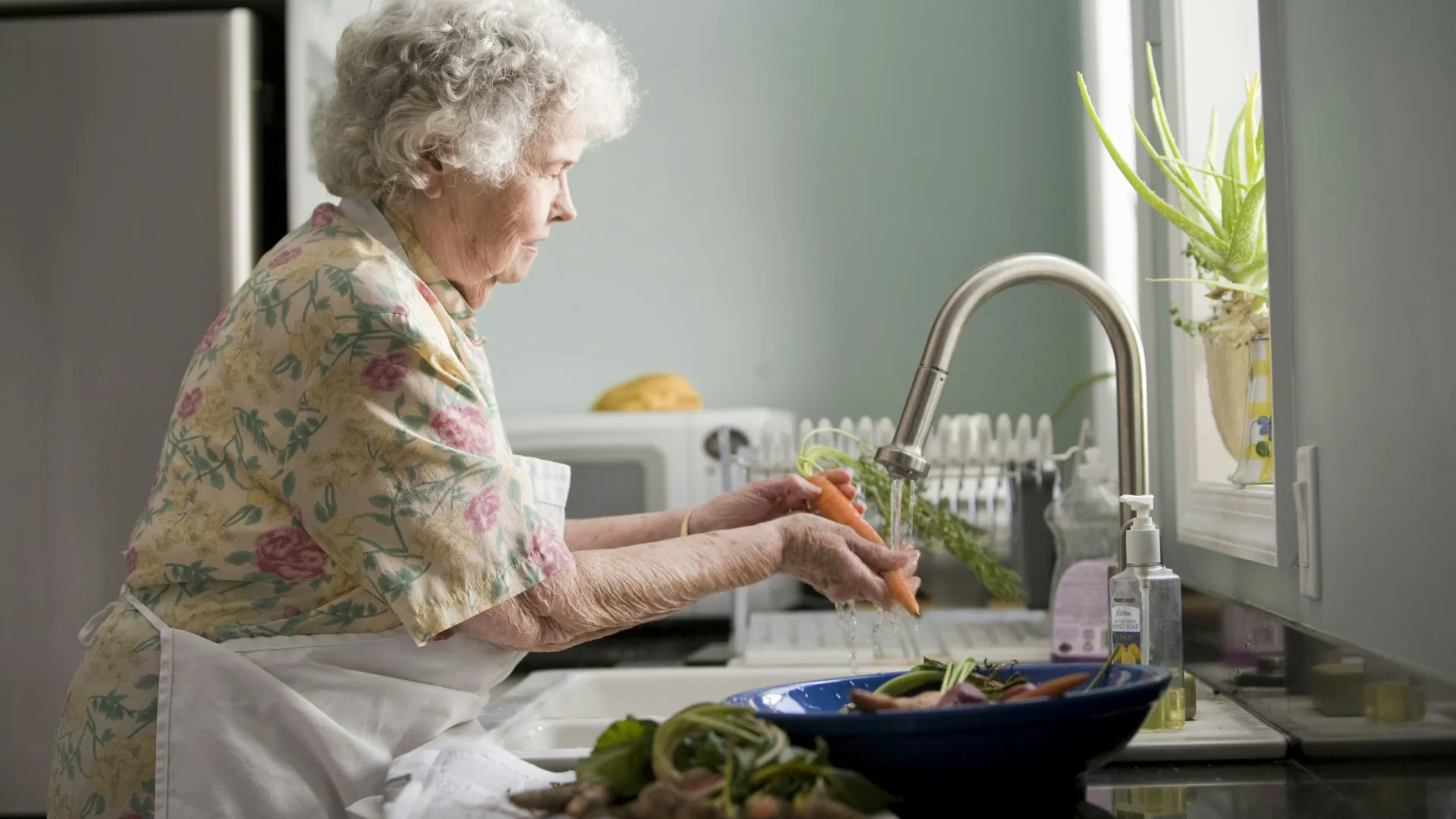 Elderly woman washing fresh carrots under kitchen sink faucet preparing vegetables for cooking.