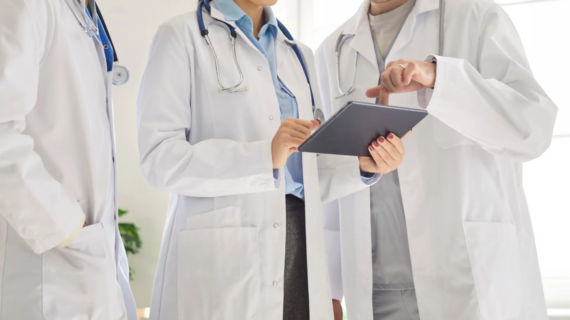 Three doctors in white coats discussing medical information using a digital tablet in a bright office.