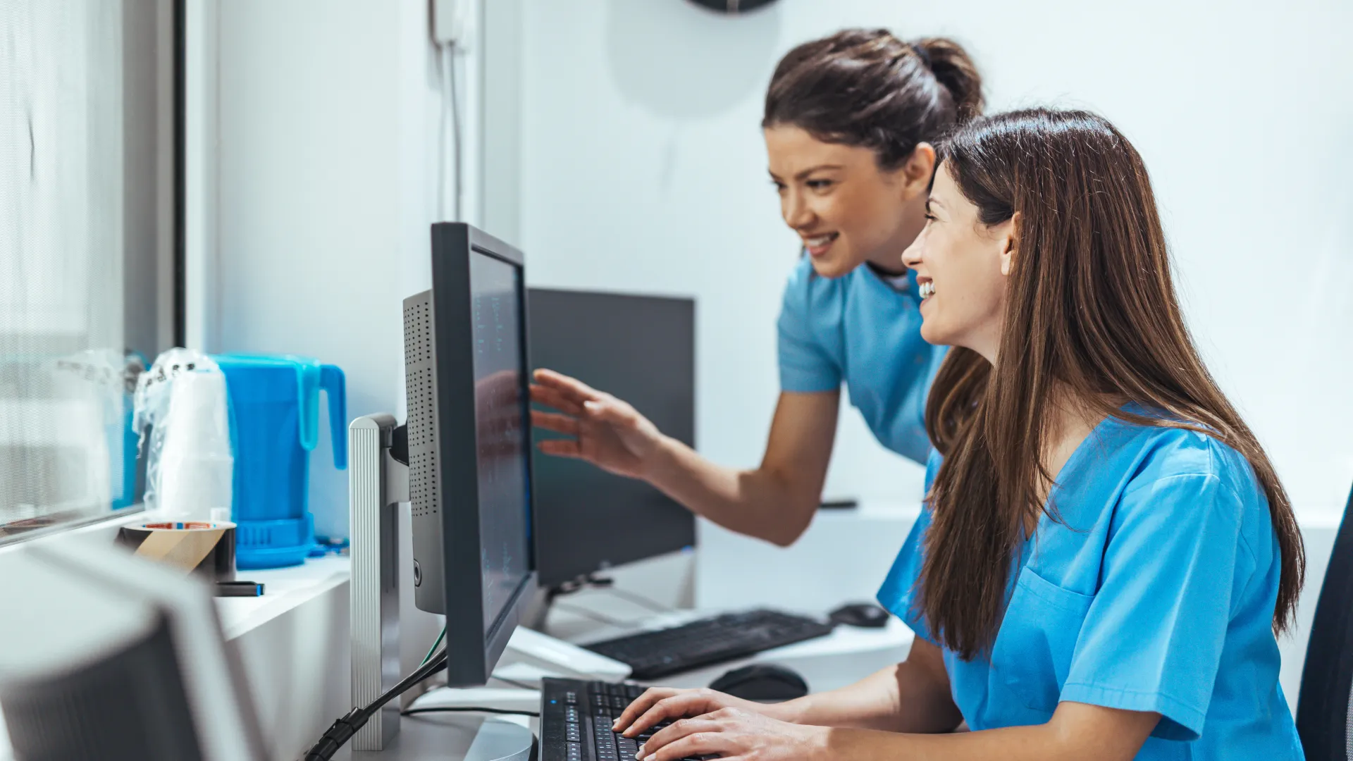 Two female healthcare professionals in blue scrubs working together on a computer in a medical office.